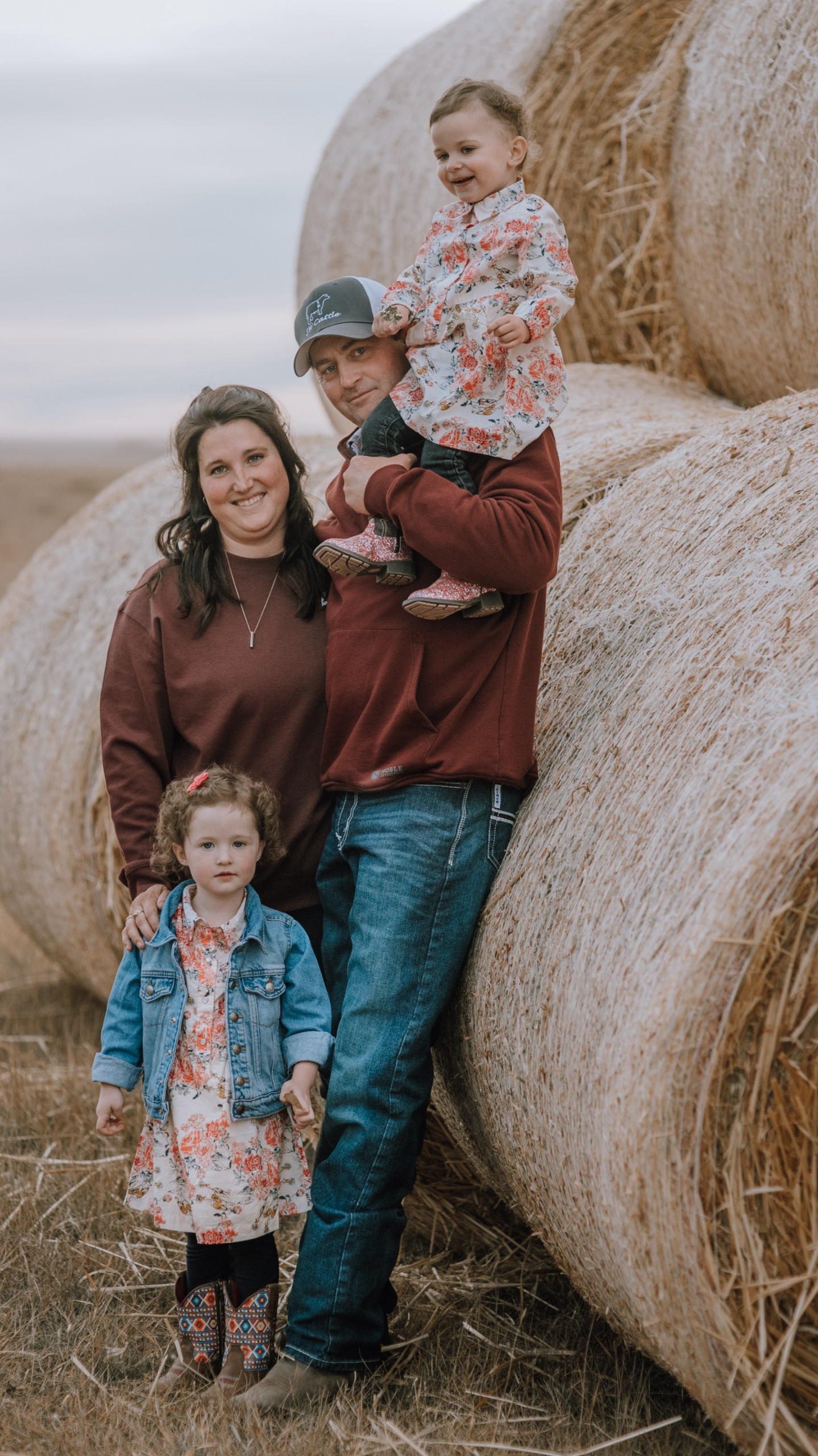 Family Picture – Wealth On Main Street Family of four posing by a large round hay bale in a rural field, smiling at the camera.